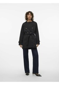 Black trench coat with double-breasted buttons, a belted waist, and wide sleeves, paired with dark denim trousers and pointed black shoes.