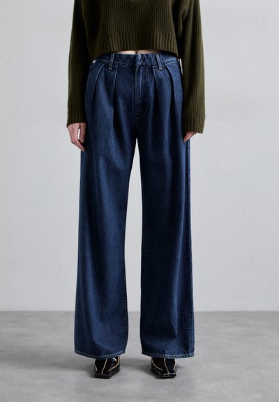 Jean en denim bleu foncé à jambes larges avec des plis à l'avant. Ils présentent une taille haute et une finition soignée, assortis à des chaussures noires.