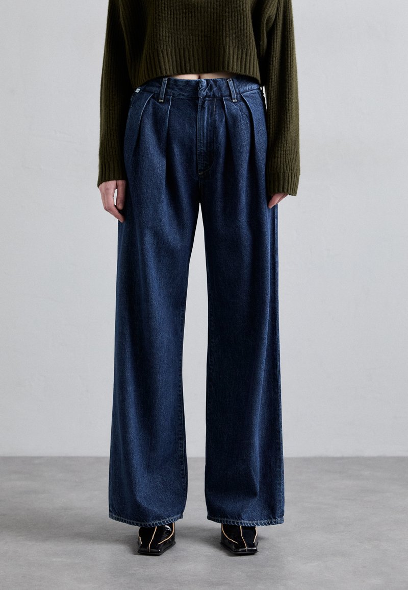 Bredde ben mørkeblå denimjeans med læg foran. De har høj talje og en ren finish, paret med sorte sko.