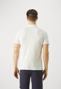 Weißes Polohemd mit kurzen Ärmeln, aus weichem Baumwollmaterial und mit einem Kragen. Verfügt über eine Rückennaht und einen figurschmeichelnden Schnitt. Wird mit dunklen Shorts getragen.
