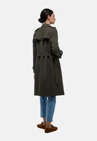 Manteau long à carreaux en vert foncé et noir, avec une taille ceinturée, des épaulettes et un détail froncé dans le dos. Porté sur un jean bleu.