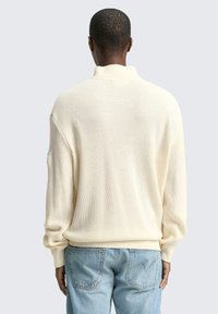 Cremfarbener Strickpullover mit hohem Kragen, gerippter Textur und abgesenkten Schulternähten, kombiniert mit hellblauen Jeans.