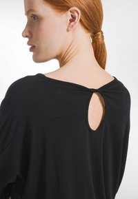 Vrouw met rood haar in een lage paardenstaart, gekleed in een zwart shirt met een kleine sleutelgatopening en een knoopdetail op de bovenrug.