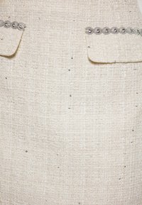 Il tessuto tweed color crema presenta una superficie textured con accenti di filo metallico e due tasche applicate, decorate con accessori argentati ornamentali.