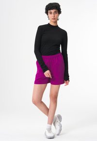 Schwarzes Langarmoberteil kombiniert mit lebhaften fuchsiafarbenen Shorts mit elastischem Bund. Getragen mit hellgrauen Sportschuhen, vor einem schlichten Hintergrund.