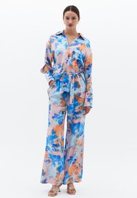 Kleurrijke tie-dye outfit met een overhemd met kraag en wijde broek in blauwe, oranje en roze tinten, gemaakt van een gladde, zijdezachte stof.