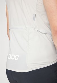 Haut sans manches gris clair avec une texture lisse, featuring des poches zippées et un logo imprimé à l'ourlet. Coupe ajustée pour le confort.
