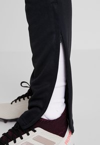 Zwarte sportbroek met ritsen aan de zijkant, met een witte sok en crèmekleurige en rode voetbalschoenen met een gestructureerd ontwerp en logo-accenten.