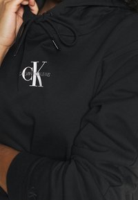 Sudadera negra con capucha y cordón, que presenta un logo blanco de "CK". Fabricada en un tejido suave, incluye puños acanalados y un corte relajado.