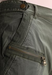 Gröna bomullsbyxor har en texturerad rem, mässingsknappar och en sidoficka med dragkedja. Tyget har en något skrynklig framtoning.