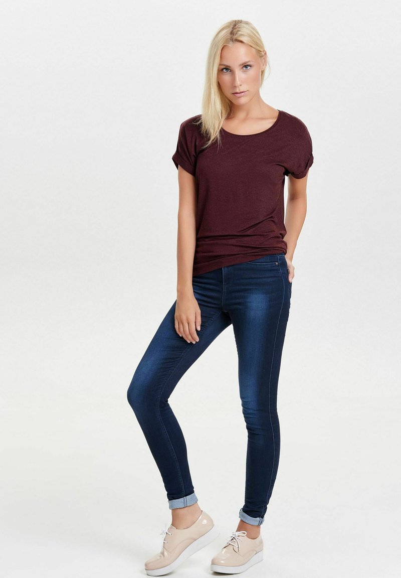 Burgunderfarget t-skjorte med oppbrettede ermer, kombinert med mørkeblå skinny jeans og lyse beige sneakers med hvite såler. Enkelt bakgrunn.