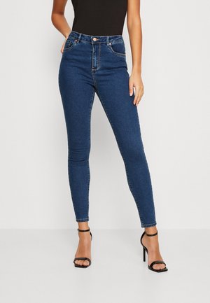 Femme portant un jean skinny bleu foncé et des sandales noires à talons hauts, debout devant un fond clair uni.