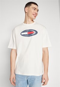 Camiseta blanca de algodón con un logo redondo en azul marino y rojo que dice "TOMMY JEANS" en el centro. Corte relajado y mangas cortas, combinada con jeans de un azul claro.