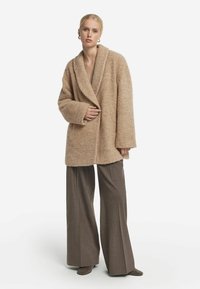 Texturerad, beige ullblandad kappa med bred krage och oversized passform. Har sänkta axlar och långa ärmar. Kombinationen med bruna vida byxor.