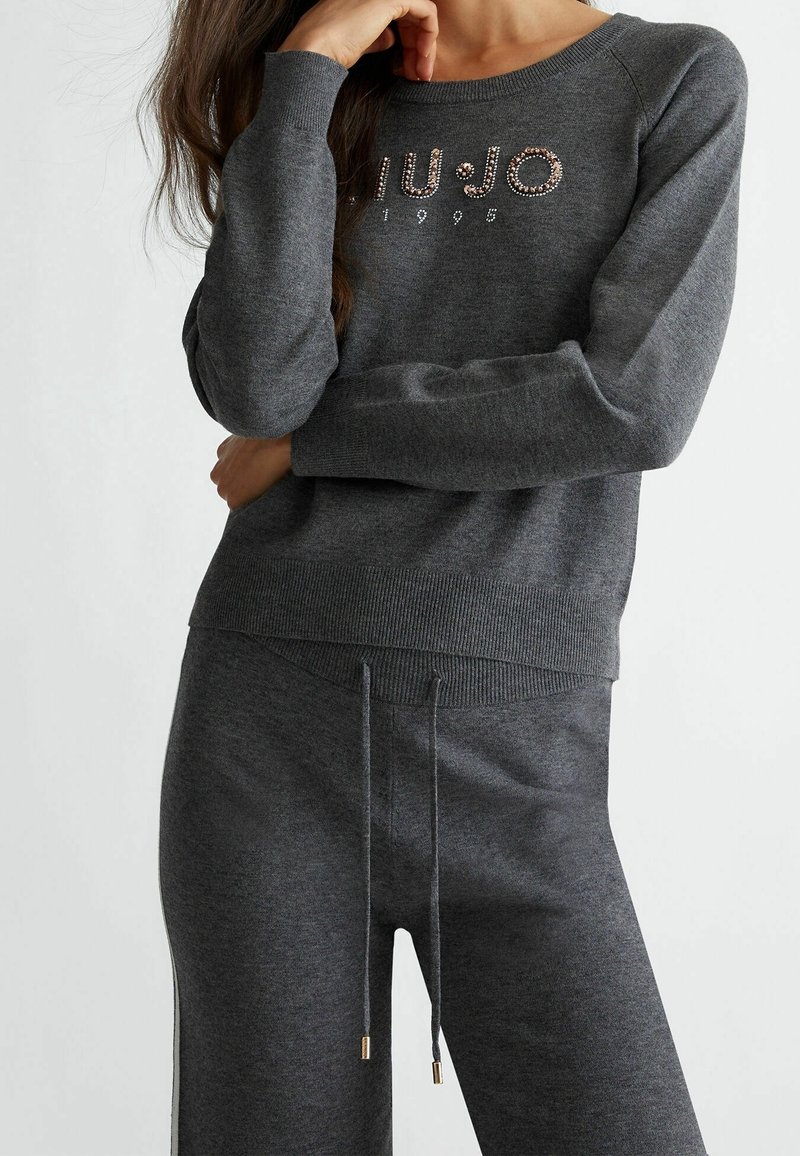 Femme portant un haut à manches longues gris foncé et un pantalon assorti, le haut orné de "LIU·JO 1995" en strass.
