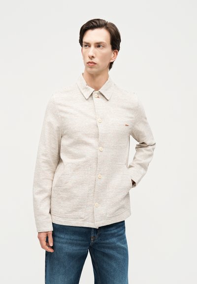 Νεαρός άνδρας με κοντά σκούρα μαλλιά, φοράει ένα ανοιχτό μπεζ υφασμάτινο σακάκι με κουμπιά και μπλε τζιν, στέκεται με το ένα χέρι στην τσέπη.