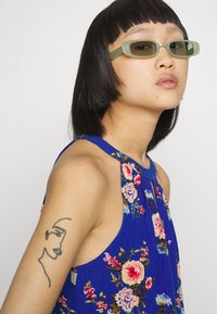 Óculos de sol verdes em formato retangular, blusa sem mangas azul com padrão floral, textura de tecido suave, cabelo curto e escuro, e uma tatuagem minimalista no braço.