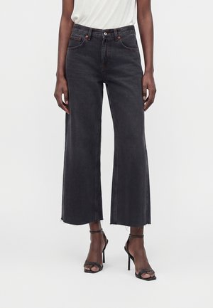 Personne portant un jean noir taille haute à jambes larges avec ourlet brut, associé à des sandales noires à talons hauts et bouts ouverts et un top blanc.