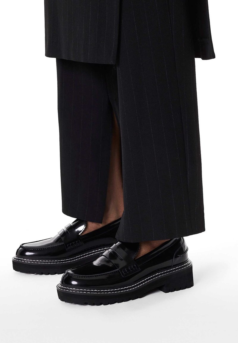 Czarne lakierowane mokasyny na masywnej podeszwie, z dekoracyjnym paskiem i kontrastującym białym przeszyciem, zestawione z spodniami w prążki.