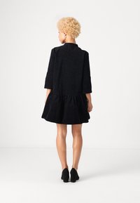 Melnas kleitas ar teksturētu leopardu rakstu, trīs ceturtdaļu piedurknēm un izplestu volānu apakšu. Uzvilkta ar melniem papēžiem, skatīta no aizmugures.