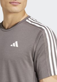 Camiseta deportiva gris con rayas de acento blancas en las mangas y un logo blanco en el pecho. Textura suave con un cuello redondo.