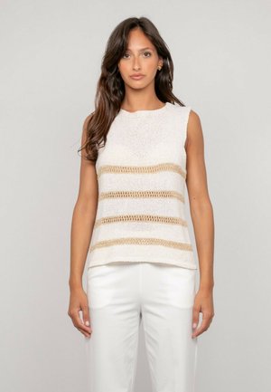 Femme aux longs cheveux bruns portant un haut en maille blanc sans manches avec des rayures horizontales beige et un pantalon blanc, debout devant un fond clair.