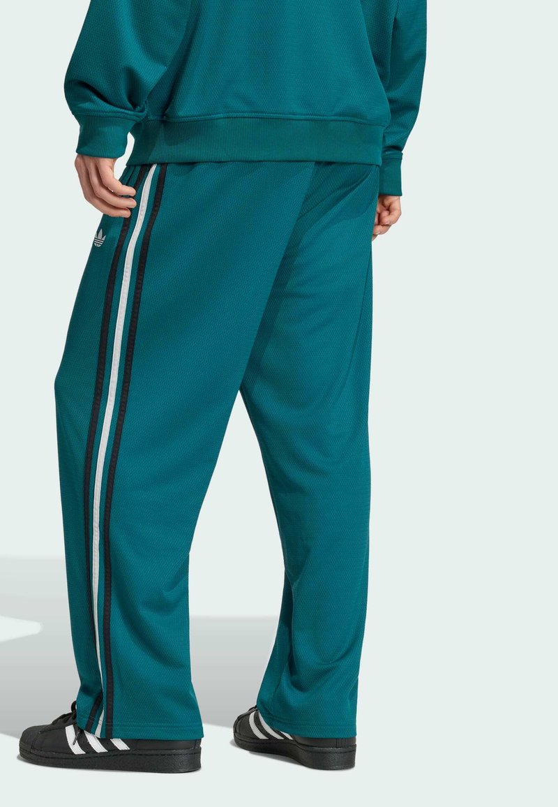 Person trägt türkisfarbene Jogginghose mit schwarzen und weißen Seitenstreifen, schwarze Sneakers mit weißen Streifen und einen passenden türkisfarbenen Pullover.