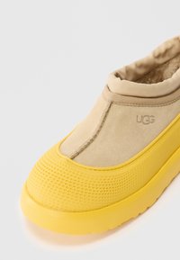 Béžové semišové slip-on boty UGG s polstrovaným límcem a jasně žlutou texturovanou gumovou podrážkou a ochranou špičky.