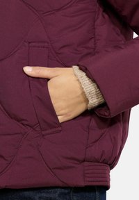 Burgundinė pūkinė striukė su siuvinėta tekstūra, turinti šoninį kišenę ir guminėmis rankovėmis, po ja uždėtas kreminės spalvos megztinis.