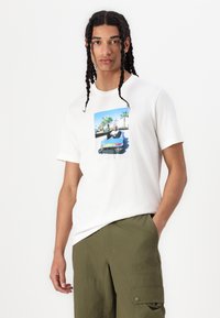 Tricou alb din bumbac cu un grafic imprimat central reprezentând o persoană într-un vehicul albastru, pe fundal cu palmieri. Este asortat cu pantaloni verzi măslinii.
