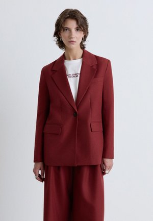Jeune personne aux cheveux courts portant un blazer rouge foncé et un pantalon à jambes larges par-dessus une chemise blanche avec un texte, debout devant un fond clair uni.