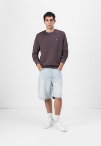 Crewneck sweatshirt i mørk lilla med gul logo, kombinert med lyseblå denimshorts, hvite sneakers og grå sokker. Minimalistisk design.