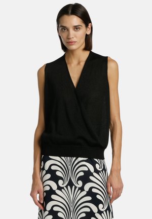 Rövid sötét hajú nő, fekete ujjatlan V-nyakú felsőt és fekete-fehér mintás szoknyát visel, egyszínű háttér előtt áll.