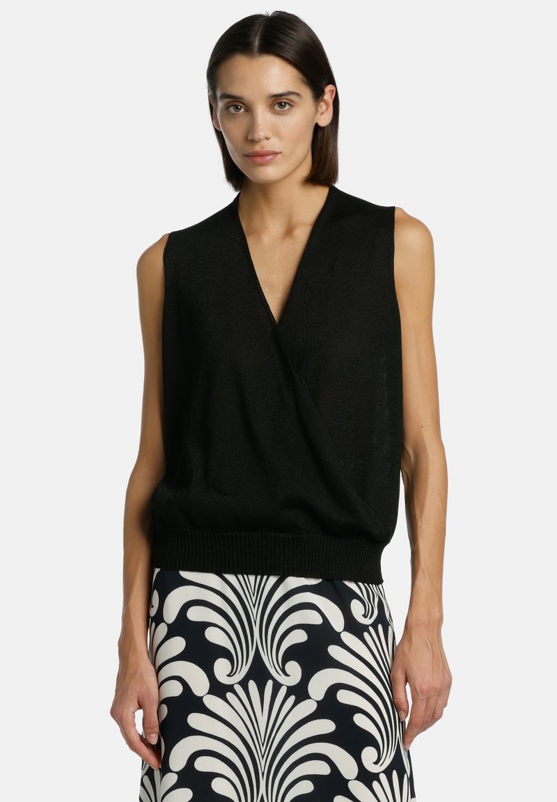 Femme aux cheveux courts et foncés portant un haut noir sans manches à col en V et une jupe à motifs noir et blanc, debout devant un fond uni.