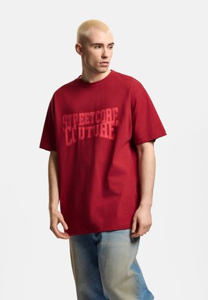 Młody mężczyzna z krótkimi blond włosami, w czerwonym t-shirtcie "Streetcore Couture" i jasnoniebieskich dżinsach, patrzący w lewo.