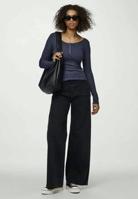 Tumši zilas garām piedurknēm henley krekls, apvienots ar melniem platām biksēm, tur melnu pleca soma, valkā melnas saulesbrilles un kedas.