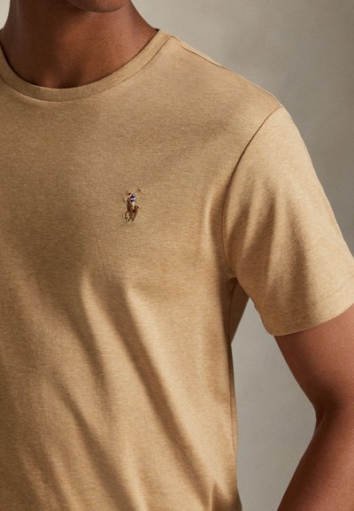 Mężczyzna noszący beżowy t-shirt z krótkim rękawem z małym haftowanym logo polo na koniu na piersi.