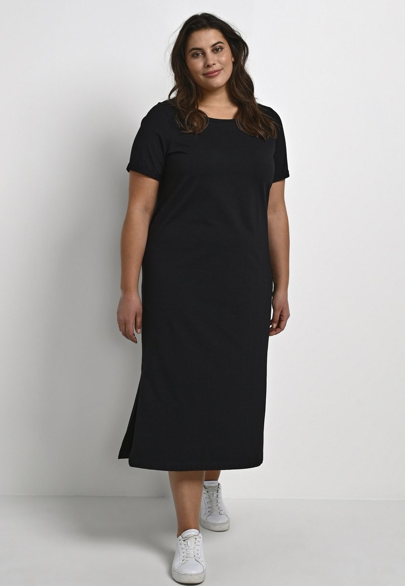 Vestido negro de algodón hasta la rodilla, con mangas cortas, cuello redondo, abertura lateral, combinado con zapatillas blancas sobre un fondo liso.