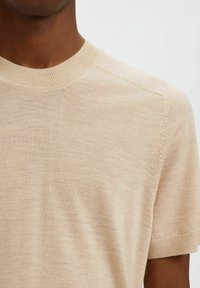 Pull en tricot beige à manches courtes avec un col rond, texture lisse et détails de couture subtils le long de l'épaule.