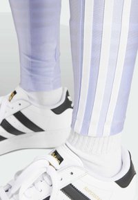 Närbild av fötter i vita Adidas Superstar-sneakers med svarta ränder, vita strumpor och ljuslila byxor med vita sidsränder.