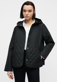 Γυναίκα με ίσια σκούρα μαλλιά, φορώντας μαύρο μπουφάν με κουκούλα και επένδυση πάνω από λευκό πουκάμισο, με το ένα χέρι στην τσέπη του μπουφάν.