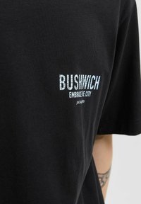 Fekete pamut póló, laza szabással. A elején kék és fehér szöveg szerepel: "BUSHWICH EMBRACE THE CITY," alatta egy logóval.