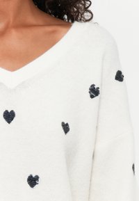 Maglione bianco con scollatura a V e decorazioni a forma di cuore in perline nere sparse. Tessuto morbido con un design a vestibilità ampia.