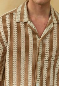 Brązowa i kremowa koszula z krótkim rękawem o geometrycznym wzorze, z zapięciem na guziki, kołnierzem z wycięciami i teksturowaną tkaniną.