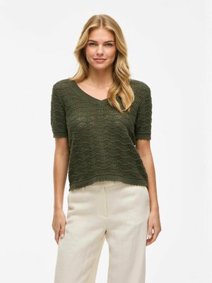 Green, short-sleeve knitted top with a V-neck, featuring a wave pattern and scalloped hem. Worn with light beige, loose-fitting pants.