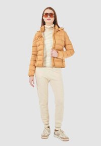 Chaqueta acolchada de caramelo con capucha, suéter blanco texturizado, pantalones de chándal beige claro y zapatillas beige con acentos metálicos.