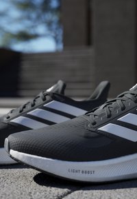 Chaussures de sport noires avec un dessus texturé, design à trois bandes blanches et une semelle blanche portant la marque "LIGHT BOOST".