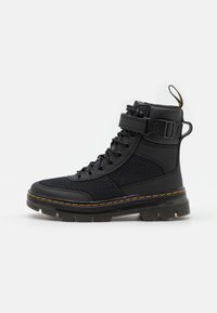 Bottes noires à la cheville en matériau synthétique lisse et en mesh, dotées d'un col rembourré, d'un système de laçage à l'avant, d'une sangle ajustable et de coutures jaunes.