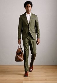 Homme marchant en avant, portant un costume vert olive, une chemise blanche, des chaussures en cuir marron et tenant un sac en cuir marron à la main droite.