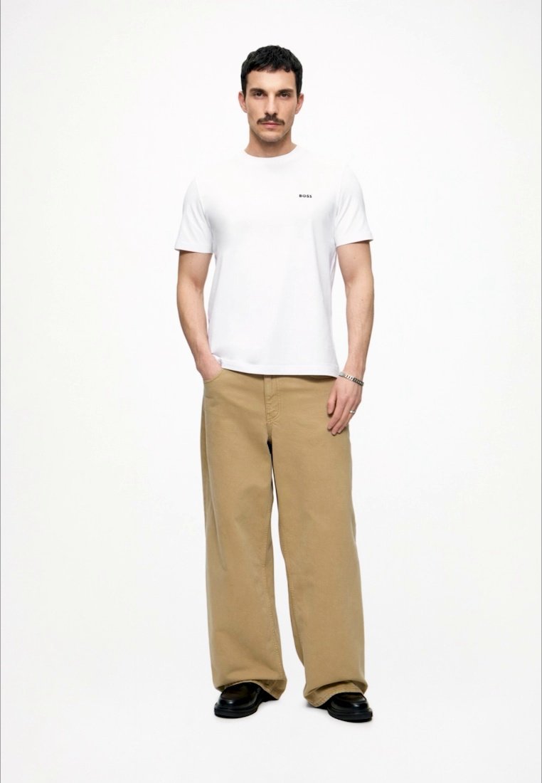 Homme debout, mains dans les poches, portant un t-shirt blanc BOSS, un pantalon ample beige, des chaussures noires et une montre-bracelet sur un fond uni.